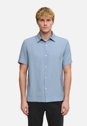 Młody mężczyzna ubrany w jasnoniebieską koszulę z krótkim rękawem i ciemne spodnie, stojący na jednolitym białym tle.