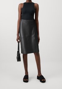 Zwarte leren midi-rok met een rechte snit en stiknaaddetails, gecombineerd met een zwarte tanktop en zwarte slides, terwijl ze een kleine handtas vasthoudt.