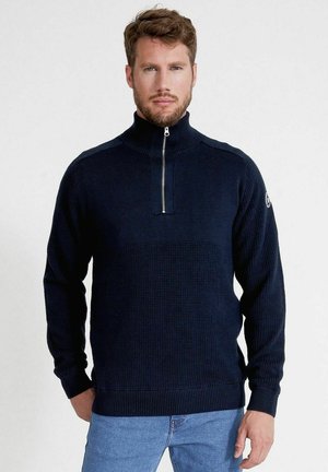 Maglione lavorato a maglia blu navy con colletto alto, mezzo zip e motivo testurizzato. Polsi e orlo a coste. Indossato con jeans azzurri chiari.