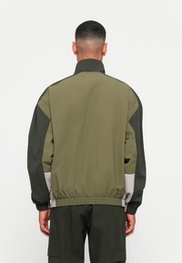 Jakna iz vetrobranega materiala v olivno zeleni barvi, ki ima visok ovratnik, elastične manšete in kontrastno sivo obrobo na robu. Narejena je iz lahkotnega materiala.