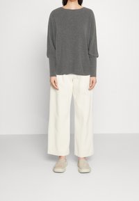 Top lungo a maniche lunghe grigio a coste con vestibilità rilassata, abbinato a pantaloni bianchi a gamba larga e scarpe beige slip-on. Design semplice e moderno.
