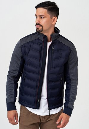 Navy puffer jacket with gray sleeves, featuring a high collar, zippered pockets, and ribbed cuffs. Smooth texture with quilted pattern.