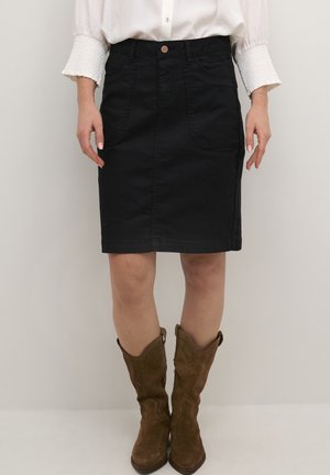 Frau trägt einen schwarzen knielangen Rock, eine weiße Bluse mit langen Ärmeln und braune Wildlederstiefel bis zur Mitte der Wade, steht vor einem einfarbigen Hintergrund.