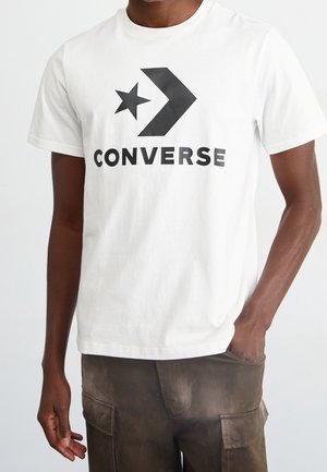 Osoba ubrana w białą koszulkę z czarnym logo Converse i brązowe spodnie typu cargo, jedna ręka w kieszeni, na jednolitym tle.