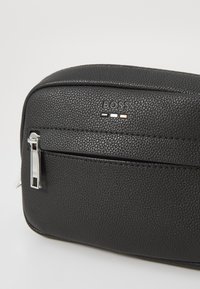 Schwarze, strukturierte Lederhandtasche mit einem silbernen Reißverschluss, versehen mit einem Logo und drei Akzentstreifen in Weiß, Grau und Roségold auf der Vorderseite.