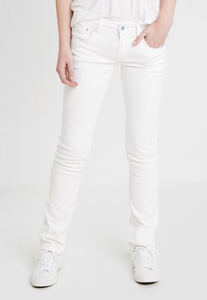 Osoba nosící bílé úzké džíny a bílé tenisky, stojící s jednou rukou v kapse před jednobarevným pozadím.