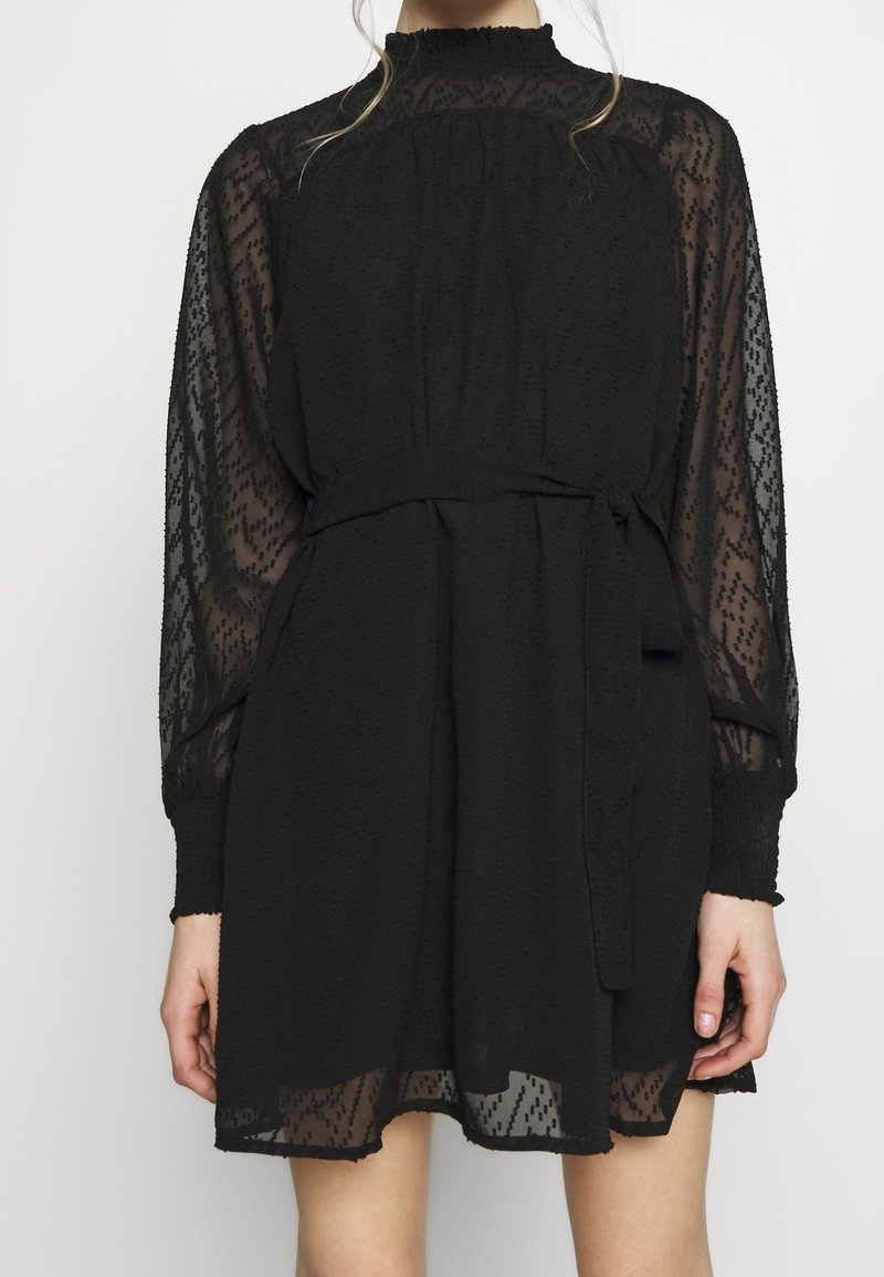 Femme portant une mini-robe noire avec des manches longues transparentes, un col montant et une ceinture nouée à la taille.