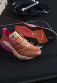 Paire de chaussures de course Adidas orange et rose avec semelles noires et blanches placées sur une surface noire avec une corde à sauter enroulée en arrière-plan.