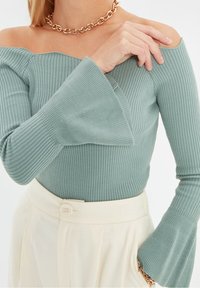 Helles grünes, geripptes Top mit schulterfreiem Design und langen, weit ausgestellten Ärmeln, kombiniert mit cremefarbenen, hoch taillierten Hosen und einer goldenen Kettenhalskette.