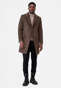 Brown wool blend double-breasted coat with large buttons, side pockets, over a light turtleneck, paired with black jeans and black lace-up boots.