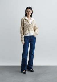 Pletený krémový pulóver s golierom, kombinovaný s tmavomodrými rozšírenými džínsami a čiernymi balerínami, na jednoduchom sivom pozadí.