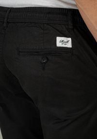 Schwarze Hose mit glatter Textur, ausgestattet mit einer Gesäßtasche mit Knopf und einem weißen Etikett, auf dem "Reell" steht. Elastischer Bund sichtbar.