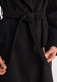 Ręce regulujące zawiązany pasek czarnego wełnianego płaszcza z bocznymi kieszeniami na osobie go noszącej.