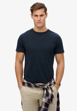 Marineblaue Rundhalsausschnitt-T-Shirt aus Baumwolle, taillierte Passform. Modell trägt hellbeige Hosen mit einem karierten Hemd, das um die Taille gebunden ist.