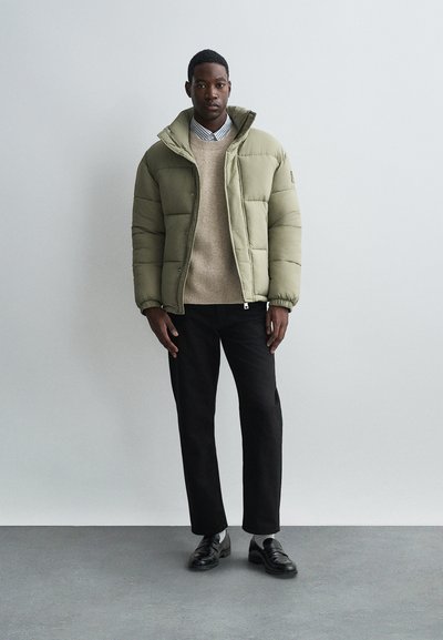 Jasnooliwkowa pikowana kurtka z wysokim kołnierzem, noszona na beżowym swetrze z dzianiny. Czarnie spodnie i czarne mokasyny dopełniają stylizację.