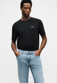 Νέος άνδρας που φοράει μαύρο T-shirt BOSS και ανοιχτόχρωμο τζιν με τα χέρια στις τσέπες, στέκεται μπροστά σε ένα απλό ανοιχτό γκρι φόντο.