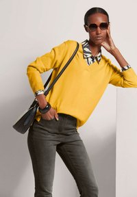 Gelber V-Ausschnitt-Pullover über einem gemusterten Hemd mit Kragen, kombiniert mit dunkelgrünen Jeans und einer kleinen schwarzen Umhängetasche. Abgerundet mit einer Sonnenbrille.