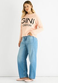 Pink fluffy sweater with black text “GINI LONDON,” paired with light blue wide-leg jeans. Model wearing pale pink ballet flats.