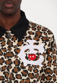 Giacca con stampa leopardata in beige e marrone con accenti neri; presenta un logo in feltro bianco con dettaglio a cuore rosso, chiusura a zip e colletto nero.