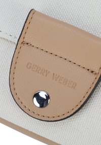 Beige synteettinen materiaali, jossa on tekstuuriset läpät, hopeanappi ja kohokuvioitu "GERRY WEBER" -logo vaaleassa kankaan taustassa.
