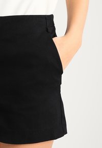 Jupe en coton mélangé noir avec une taille cintrée et des poches latérales. Elle présente une texture lisse, des lignes épurées et une longueur courte.