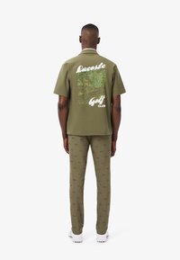 Man in olijfgroen Lacoste Golfclub shirt en bijpassende broek met kleine krokodillenlogo's, staand met zijn rug naar de camera op een witte achtergrond.