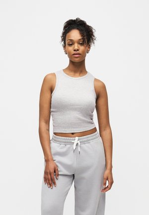 Junge Frau mit lockigem Haar, die ein hellgraues, ärmelloses Crop-Top und passende Jogginghosen trägt, vor einem weißen Hintergrund stehend.