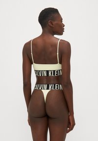 Gul stripete bralette og thong-sett med svarte "CALVIN KLEIN" logo bånd. Justerbare stropper, glatt tekstur, design med minimal dekning.