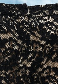 Μαύρο ύφασμα δαντέλας με λουλούδια πάνω από μπεζ φόδρα, που δείχνει λεπτομερή υφή και κυματιστή άκρη κοντά σε μια ραφή.