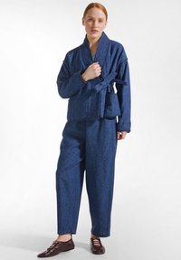 Žena s červenými vlasy, která má na sobě modrou bundu ve stylu kimona s páskem, široké kalhoty stejných barev a tmavě hnědé šněrovací boty.