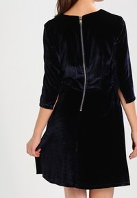 Vestido de terciopelo azul marino con mangas de tres cuartos, que cuenta con un cierre de cremallera visible en la parte posterior y un dobladillo acampanado. Textura suave y diseño limpio.