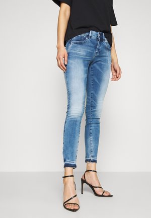 Jeans ajustados de mezclilla azul con un acabado deslavado. Cuentan con una cintura de altura media y dobladillos deshilachados. Combinados con sandalias de tacón alto negras y con tiras.