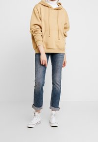 Felpa oversize beige con tasca a marsupio e cordini, abbinata a jeans azzurri arrotolati e sneakers bianche alte.