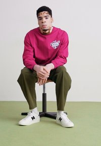 Fuksjowa bluza z długim rękawem z graficznym logo, w połączeniu z oliwkowymi spodniami i białymi trampkami z czarnymi akcentami, siedząca na stołku.