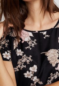 Blouse noire à fleurs avec des fleurs roses et blanches et des accents beiges. Le design a une coupe décontractée et un col décolleté.
