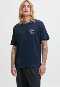 Tamsiai mėlynas medvilninis marškinėliai su apvalia iškirpte, trumpomis rankovėmis ir baltu logotipu, spausdintu kairiajame krūtinės pusėje. Lygus paviršius ir atsipalaidavęs siluetas.