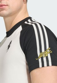 Ανδρικό εφαρμοστό t-shirt με μαύρα κοντά μανίκια, λευκό σώμα, τρεις μαύρες ρίγες στους ώμους και κίτρινο κεντητό λογότυπο στο μανίκι.