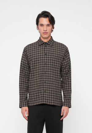 Mees, kes kannab tumepruuni ja beeži ruudulist pika varrukaga särk koos mustade pükstega, seistes ühevärvilise tausta ees.