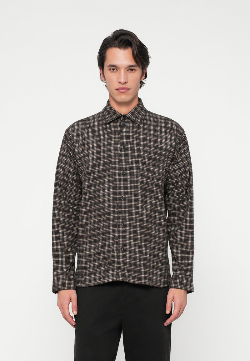 Uomo che indossa una camicia a maniche lunghe a quadri marrone scuro e beige con pantaloni neri, su uno sfondo neutro.