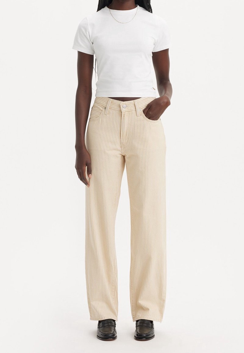 Personne noire portant un t-shirt court blanc, un pantalon large beige rayé, des mocassins noirs et un fin collier en or, debout avec une main dans la poche.