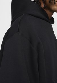 Černá mikina s kapucí, která má strukturovanou kapuci, měkký bavlněný materiál a viditelné švy na rameni. Texturovaná tkanina.