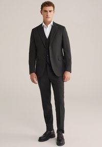 Melns trīsd auto uzvalks ar pielāgotu žaketi, vestīti un šaurām biksēm. Apvienots ar melnām kurpēm un baltu krekli.