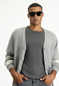 Cárdigan gris con cierre de botones, presenta acentos a rayas en gris claro y blanco en las mangas. Se lleva sobre una camiseta gris y se complementa con gafas de sol.