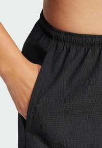 Black textured shorts with an elastic waistband and side pockets. Visible hand resting in the left pocket. Fabric has a raised pattern.