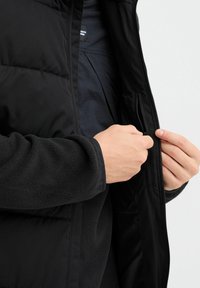 Juodas pūkinis švarkas su užtrauktuku, turintis tekstūruotą pūkinę pamušalą ir vidinį kišenę. Rankos siekianti išorinę kišenę.