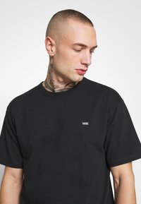 Camiseta de algodón negra con cuello redondo, mangas cortas y un pequeño logo blanco de "VANS" en la parte izquierda del pecho. Textura suave.