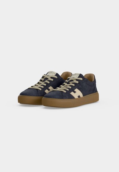 Paire de baskets bleu marine avec lacets beiges, logo latéral beige et semelles en caoutchouc marron sur fond gris clair uni.