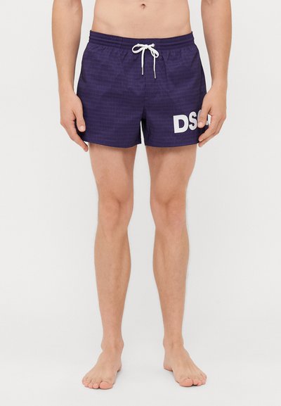 Male wearing dark purple swim shorts with white drawstring and large white "DSQ" logo on right leg, standing barefoot on plain background.