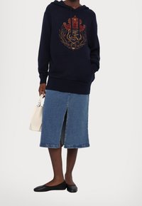 Sweat à capuche bleu marine avec poche avant et broderie dorée, associé à une jupe en denim bleu clair avec une fente à l'avant. Ballerines noires et sac blanc.