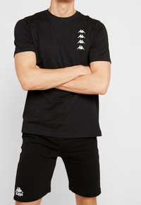 Černé triko Kappa s kulatým výstřihem, krátkými rukávy a bílými prvky loga. Spárováno s černými šortkami s bílým logem. Hladká tkanina.